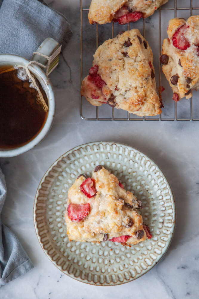 A chocolate and strawberry scone on a plate, with a mug of tea and a wire rack of more scones next to it. 