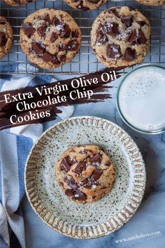 An extra virgin olive oil chocolate chip cookie on a plate, with a glass of milk next to it and more cookies on a wire rack near by.