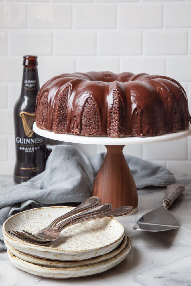 A Guinness chocolate bundt cake with Irish whiskey chocolate ganache glaze on a cake stand, with a bottle of Guinness stout beer behind it. There are small dessert plates in front of the cake with forks on the plates.