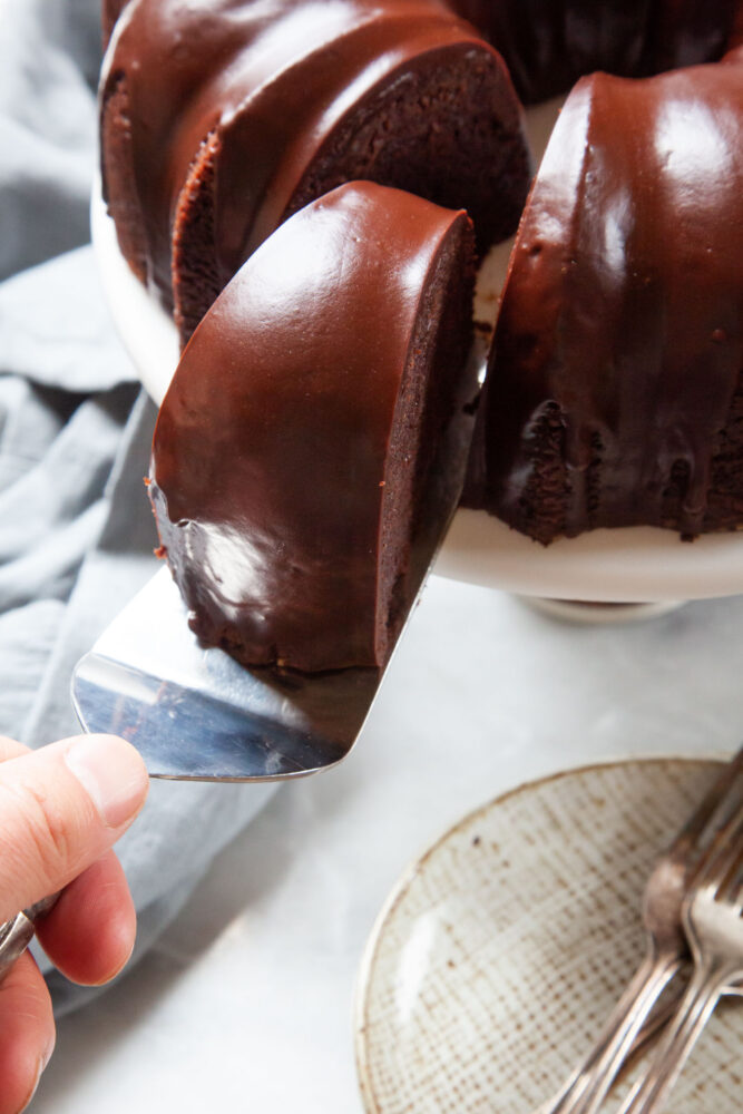 A hand holding a cake server with a slice of bundt cake on it, being pulled away from the rest of the cake on a cake stand. There are small dessert plates under the cake stand.