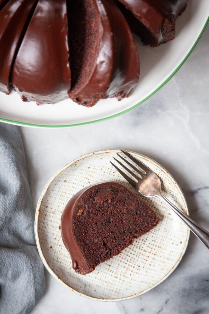 A slice of Guinness chocolate bundt cake on a plate, with a fork next to it. The remaining cake is on a cake stand next to it.