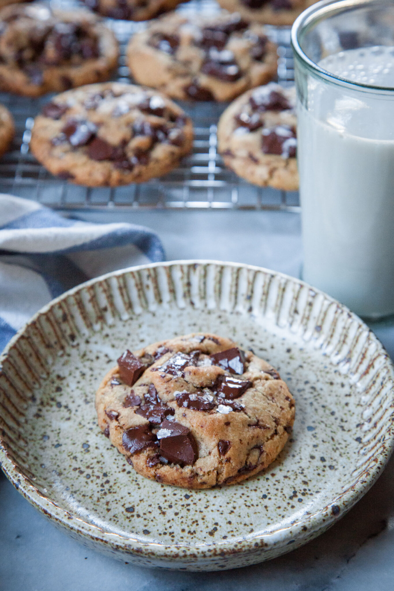 An extra virgin olive oil chocolate chip cookie on a plate, with a glass of milk next to it and more cookies on a wire rack near by.