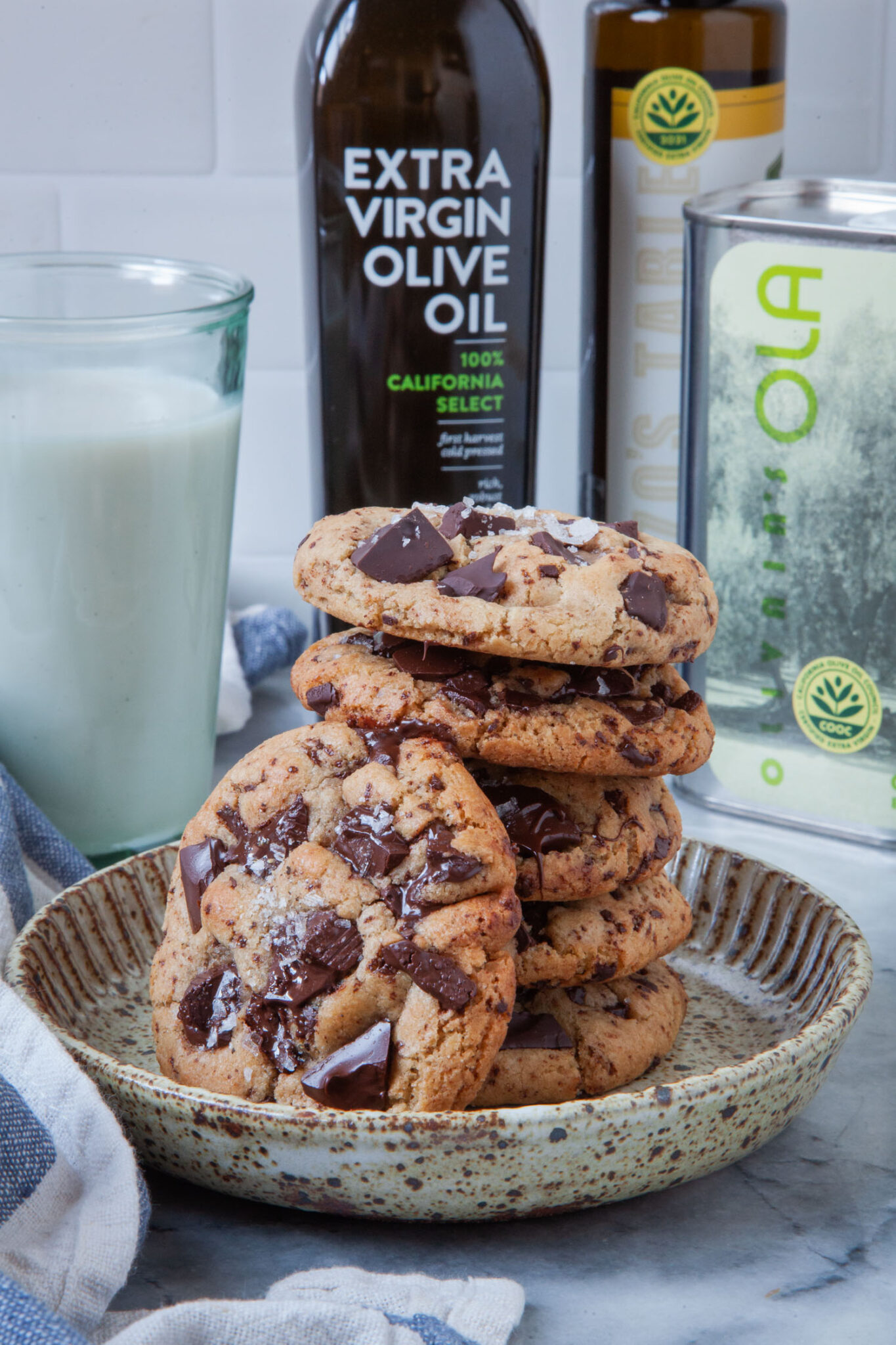 A plate with a stack of extra virgin olive oil cookies on it, with bottles of extra virgin olive oil behind it, along with a glass of milk.