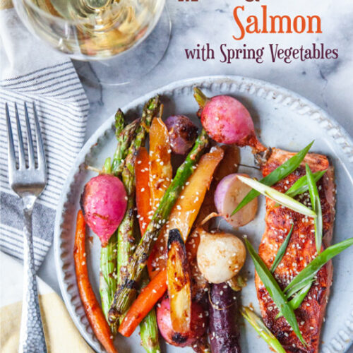 Miso-glazed salmon with a mix of roasted carrots, asparagus and radishes on a plate. There is a fork on a cloth napkin and a glass of white wine next to the plate.