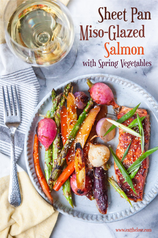 Miso-glazed salmon with a mix of roasted carrots, asparagus and radishes on a plate. There is a fork on a cloth napkin and a glass of white wine next to the plate.