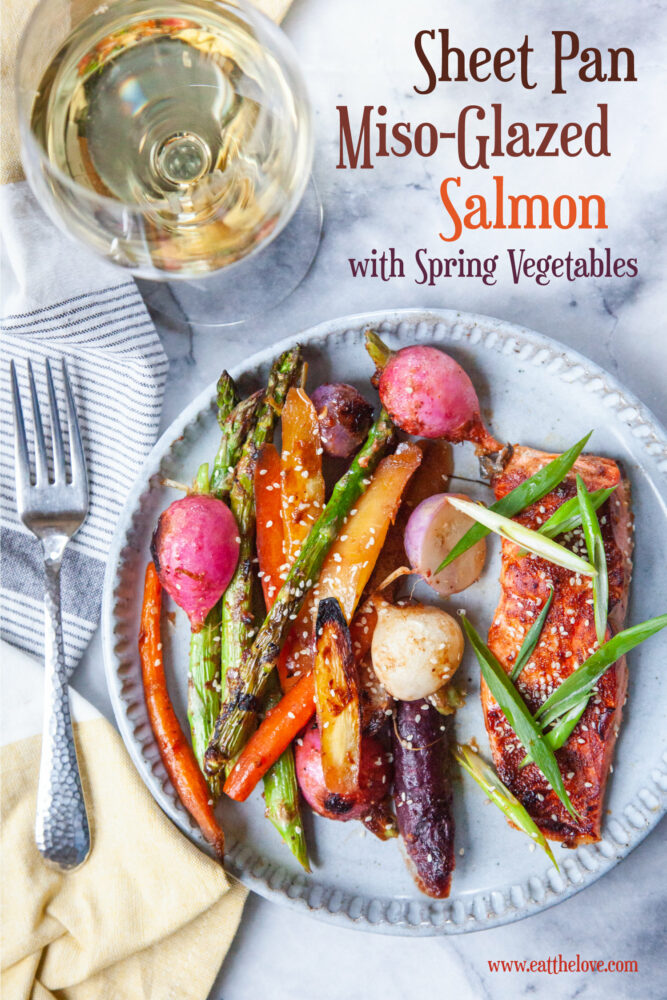 Miso-glazed salmon with a mix of roasted carrots, asparagus and radishes on a plate. There is a fork on a cloth napkin and a glass of white wine next to the plate.