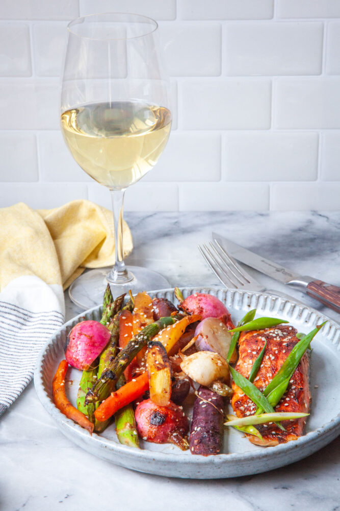 A plate of miso-glazed salmon with roasted asparagus, carrots and radishes next to it and a glass of white wine behind it.