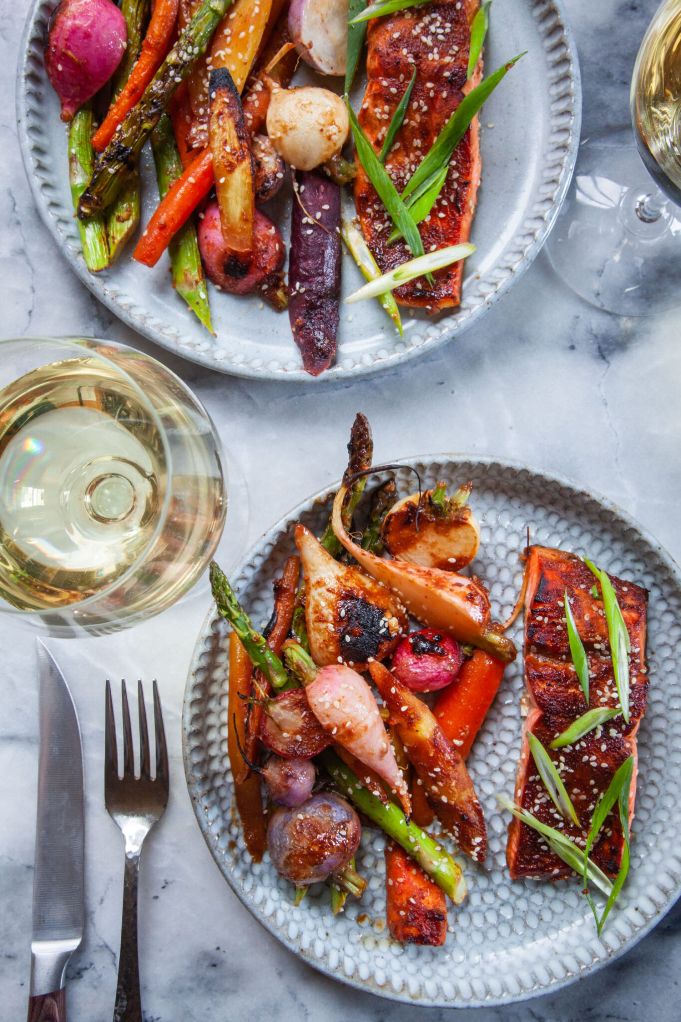 Two dinner plates with miso-glazed salmon and roasted asparagus, radishes and carrots on it. Next to the plates are glasses of white wine.