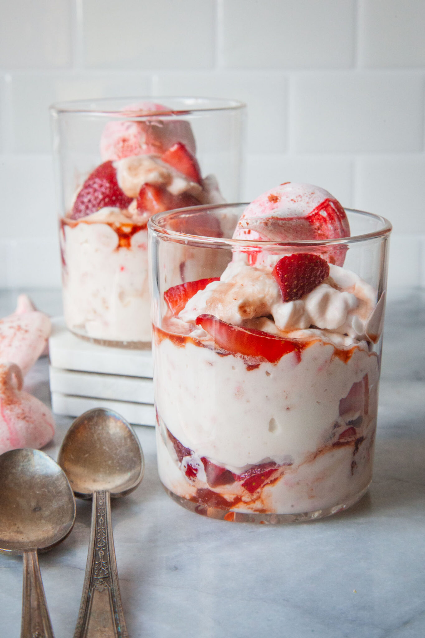 A double old fashion glass filled with British Eton Mess, a strawberry, meringue and whipped cream dessert. There is another glass of Eton Mess behind it on a marble coasters, with a spoons on the table, and a couple of meringue cookies sitting near by.