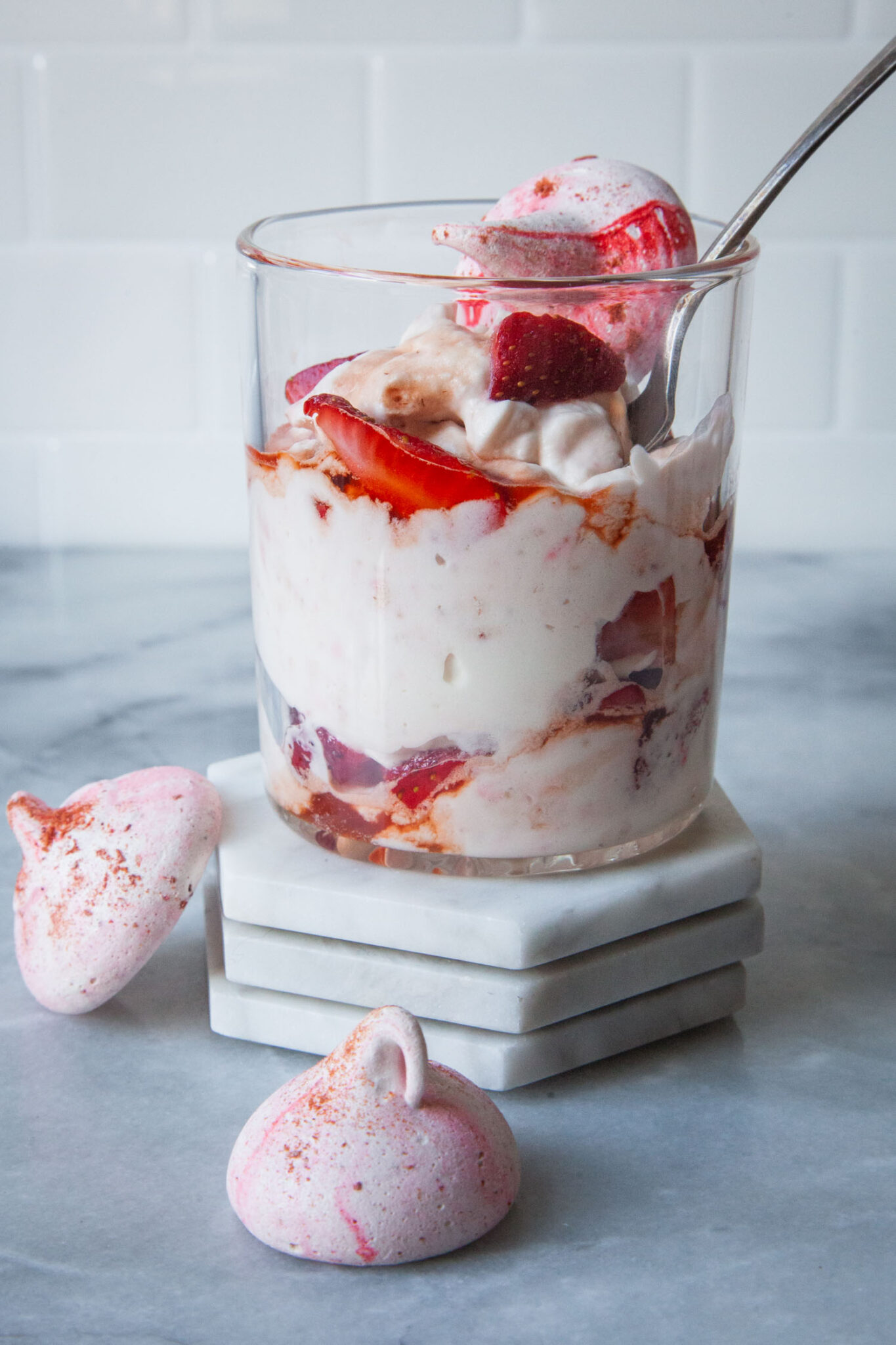 A double old fashion glass filled with British Eton Mess, a strawberry, meringue and whipped cream dessert sitting on marble coasters. There are a two meringue cookies on the table next to the Eton Mess.