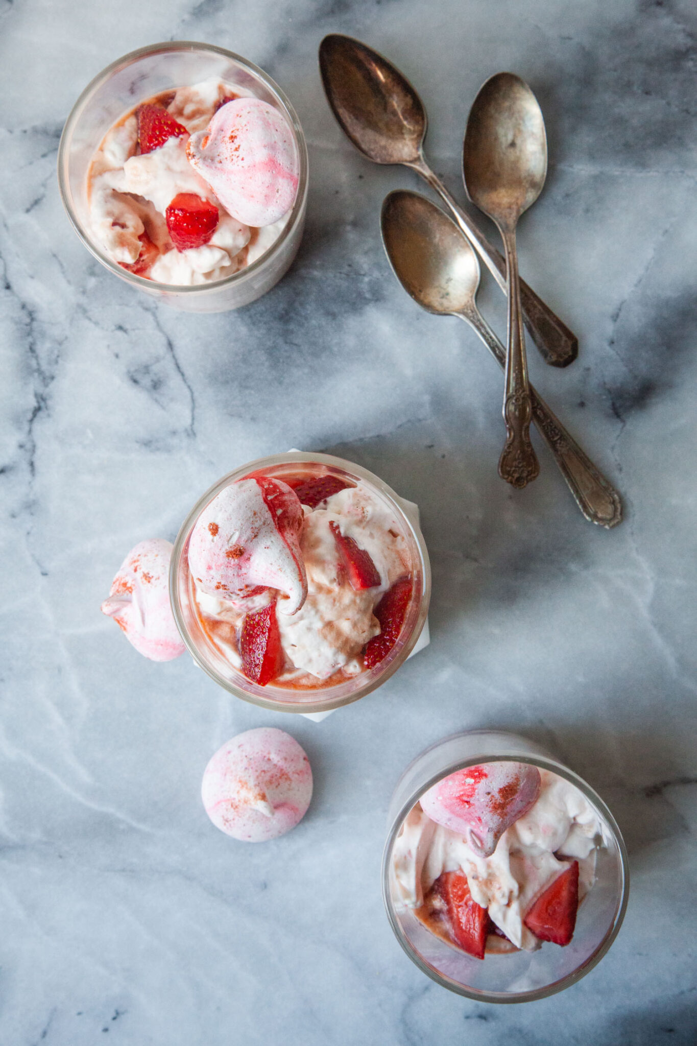 Three British Eton Mess desserts in double old fashion glasses, on a marble surface with spoons next to them. There are a couple of meringue cookies next to the glasses.