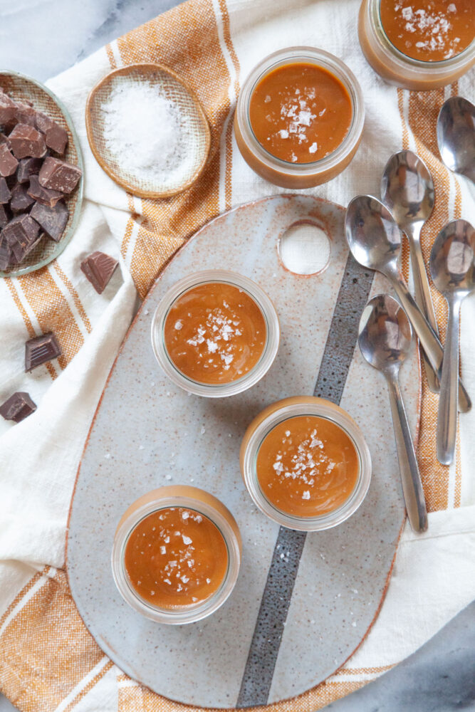 Several black bottom caramel pots de creme on a ceramic cheese board and on a cloth napkin. There are soon next to the pots de creme, along with small bowls with salt and chopped chocolate in them.