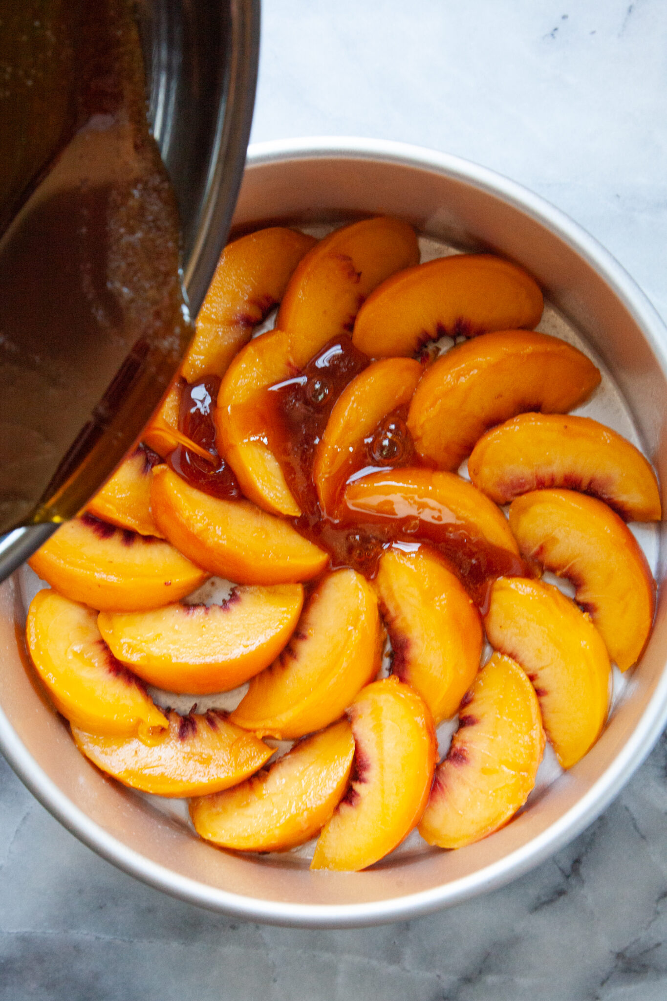 A skillet of burnt sugar caramel being poured onto slices of peaches arranged in a cake pan.
