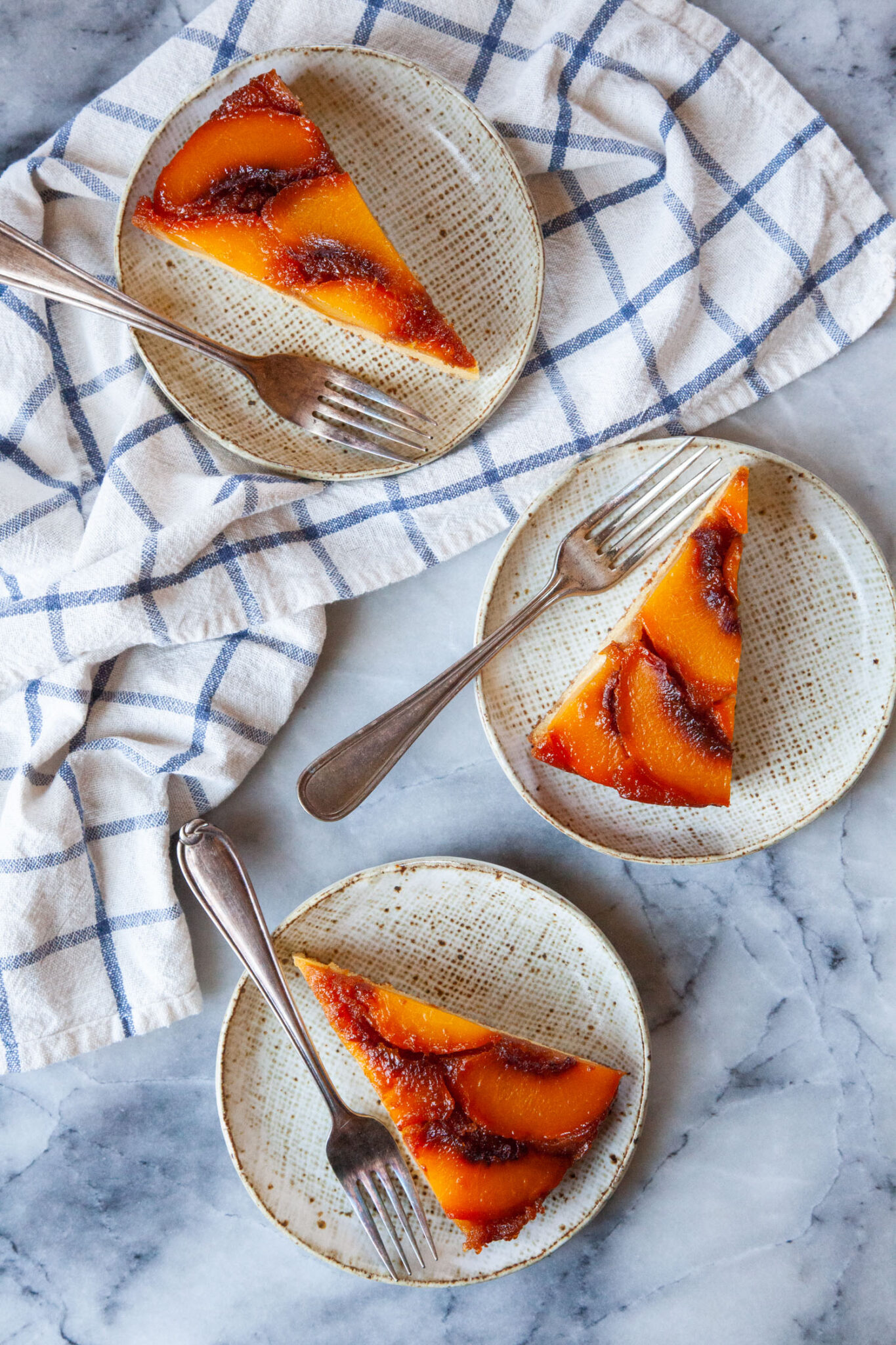 Three small plates with slices of caramel peach upside down cake on them. They each have a fork on the plate, and they are sitting next to a cloth napkin.