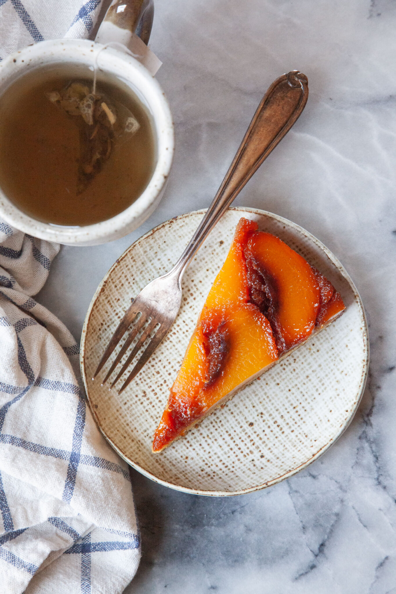 A slice of caramel peach upside down cake on a small plate, with a mug of tea next to it.