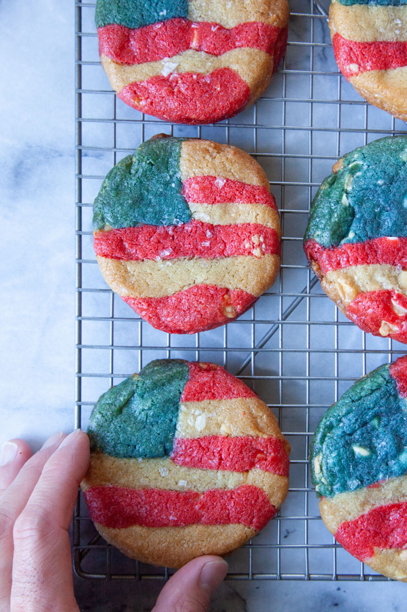 A hand reaching for a red, white, and blue American flag-inspired cookie sitting on a wire rack.