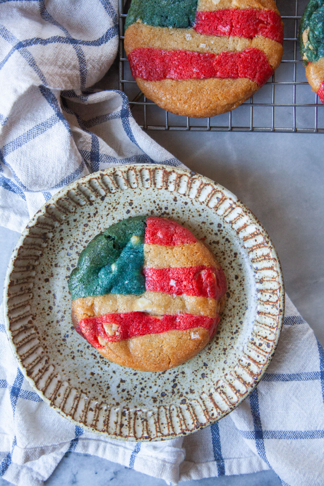 A red, white, and blue, American flag-inspired cookie sitting on a plate, with more cookies on a wire cooling rack next to it.