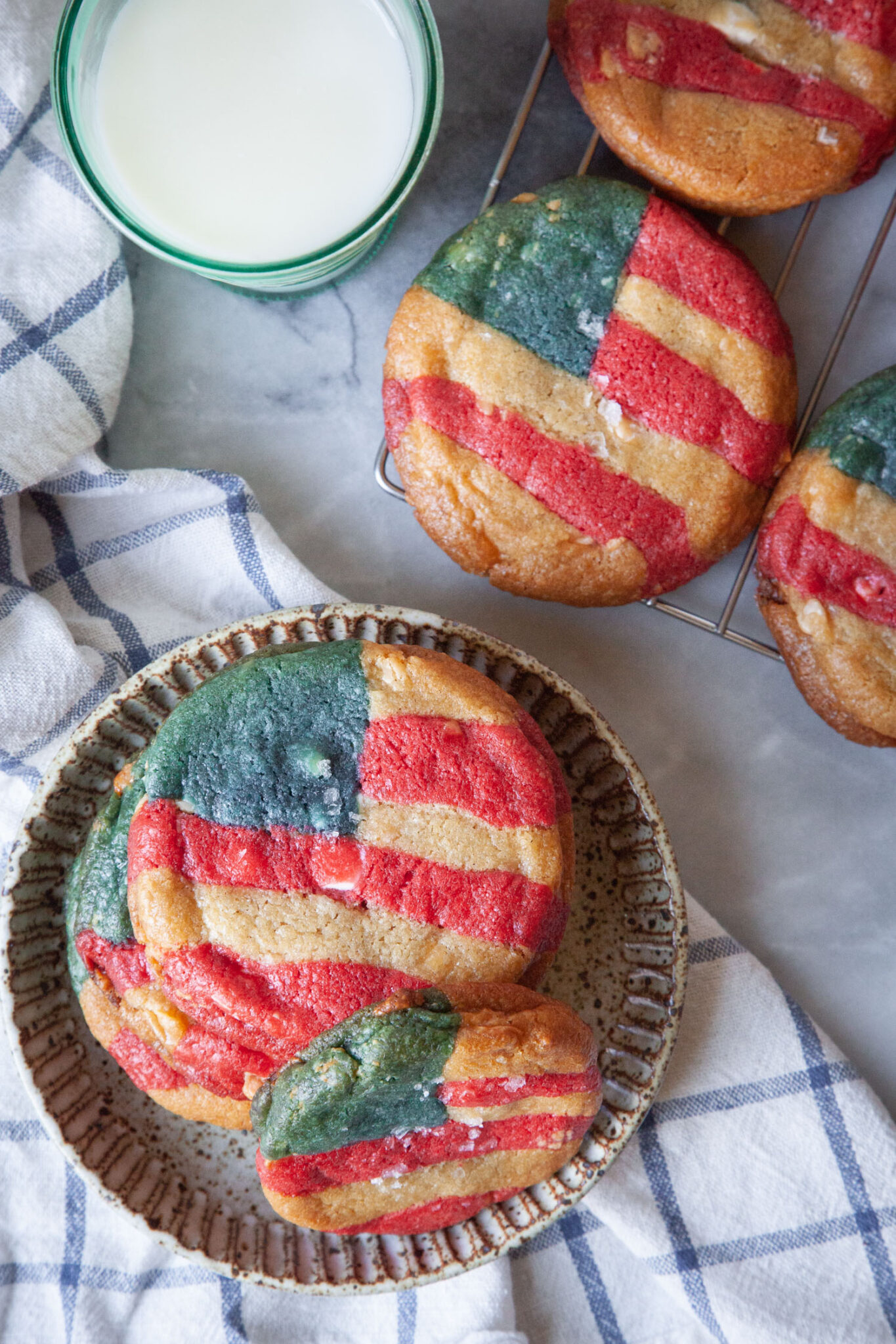 A stack of red, white, and blue American flag-inspired cookies on a plate with a glass of milk and more cookies on a wire cooling rack next to the plate.