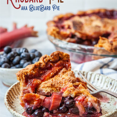 A slice of blueberry rhubarb pie on a small plate, with a bowl of blueberries and some rhubarb behind it. The remaining pie is also behind the pie slice.
