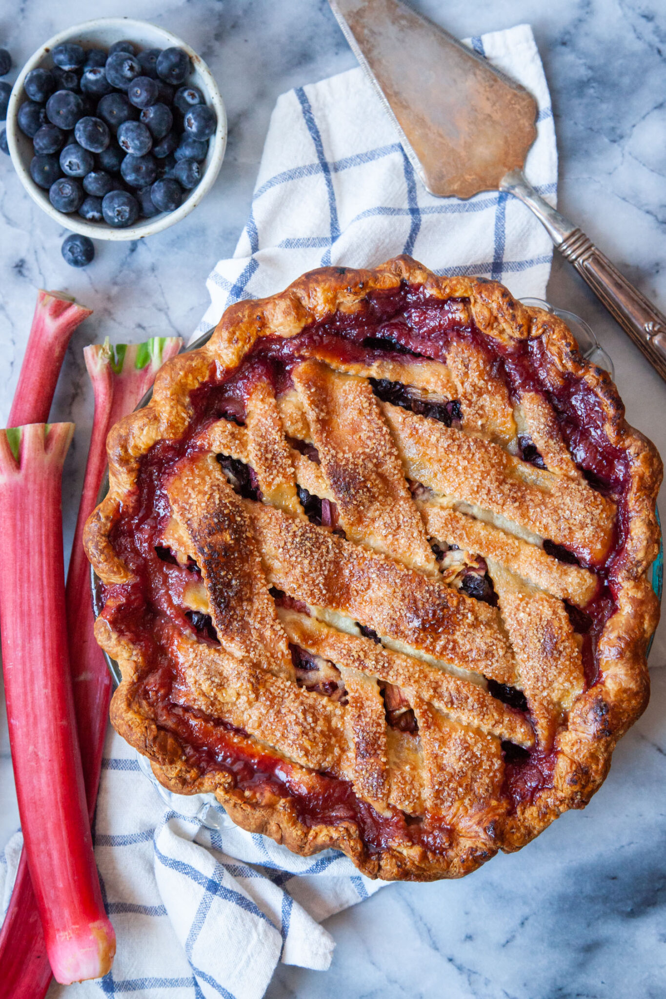 A blueberry rhubarb pie with rhubarb and a small bowl of blueberries next to it, along with a silver pie server.