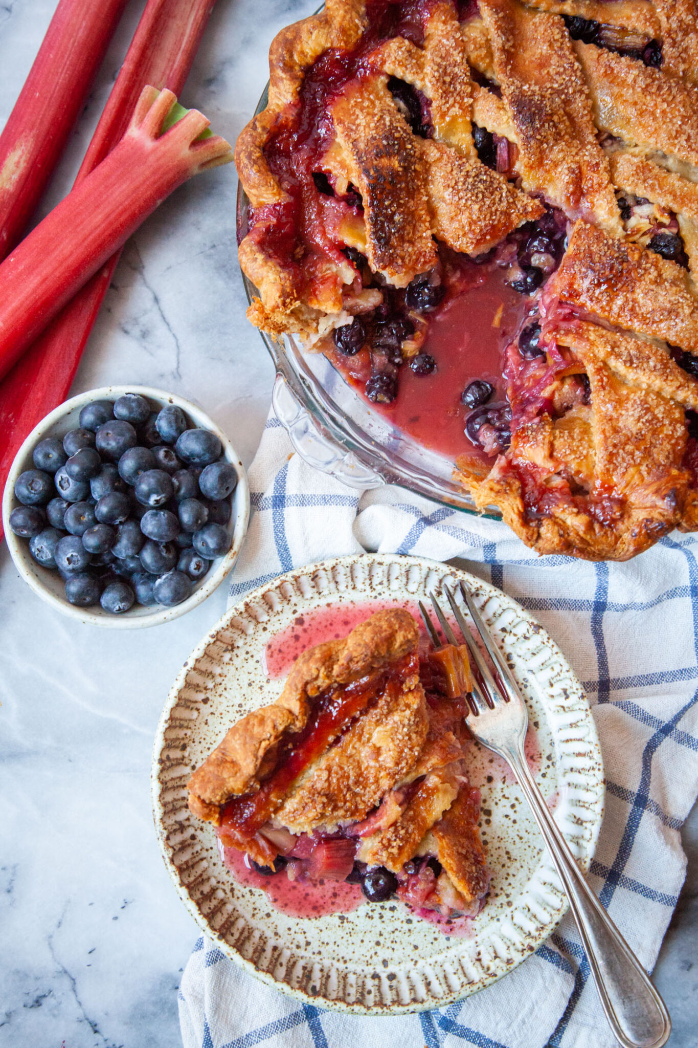 A slice of blueberry rhubarb pie on a plate, with the remaining pie next to it. Stalks of rhubarb and a small bowl of blueberries are next to the pie.
