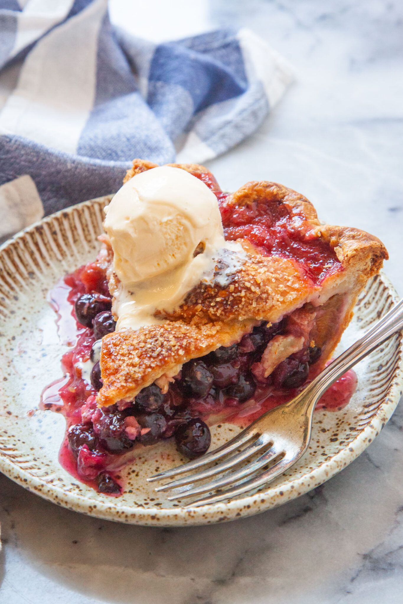 A slice of blueberry rhubarb pie on a plate, with a scoop of vanilla ice cream on it.