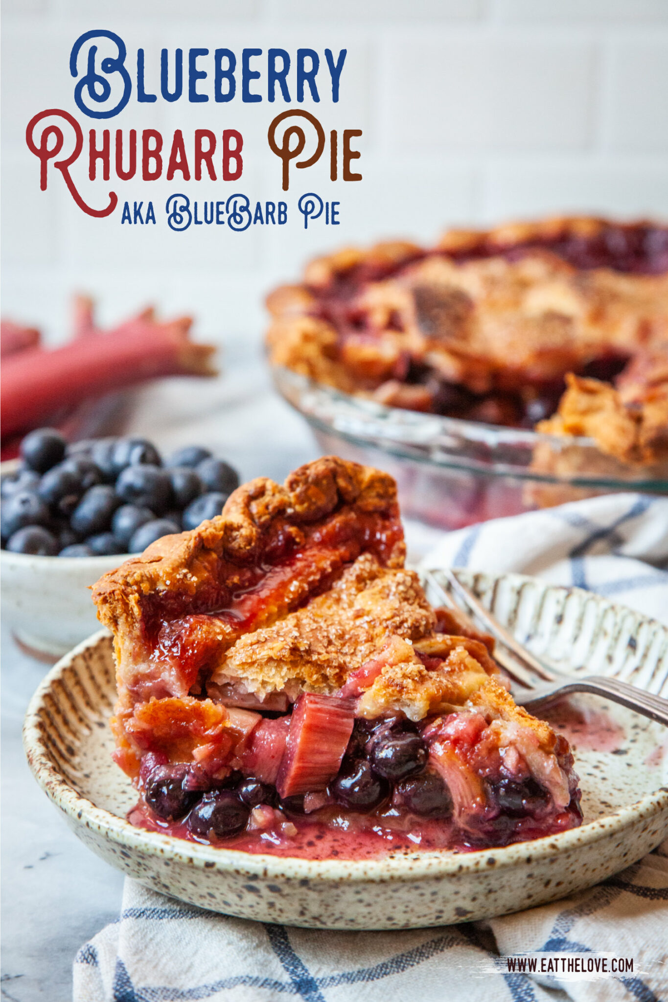 A slice of blueberry rhubarb pie on a small plate, with a bowl of blueberries and some rhubarb behind it. The remaining pie is also behind the pie slice.