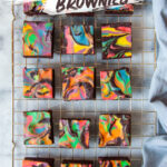 Rainbow cheesecake brownies sitting on a wire cooling rack. There is a blue gray cloth napkin next to the wire rack.