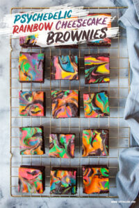 Rainbow cheesecake brownies sitting on a wire cooling rack. There is a blue gray cloth napkin next to the wire rack.