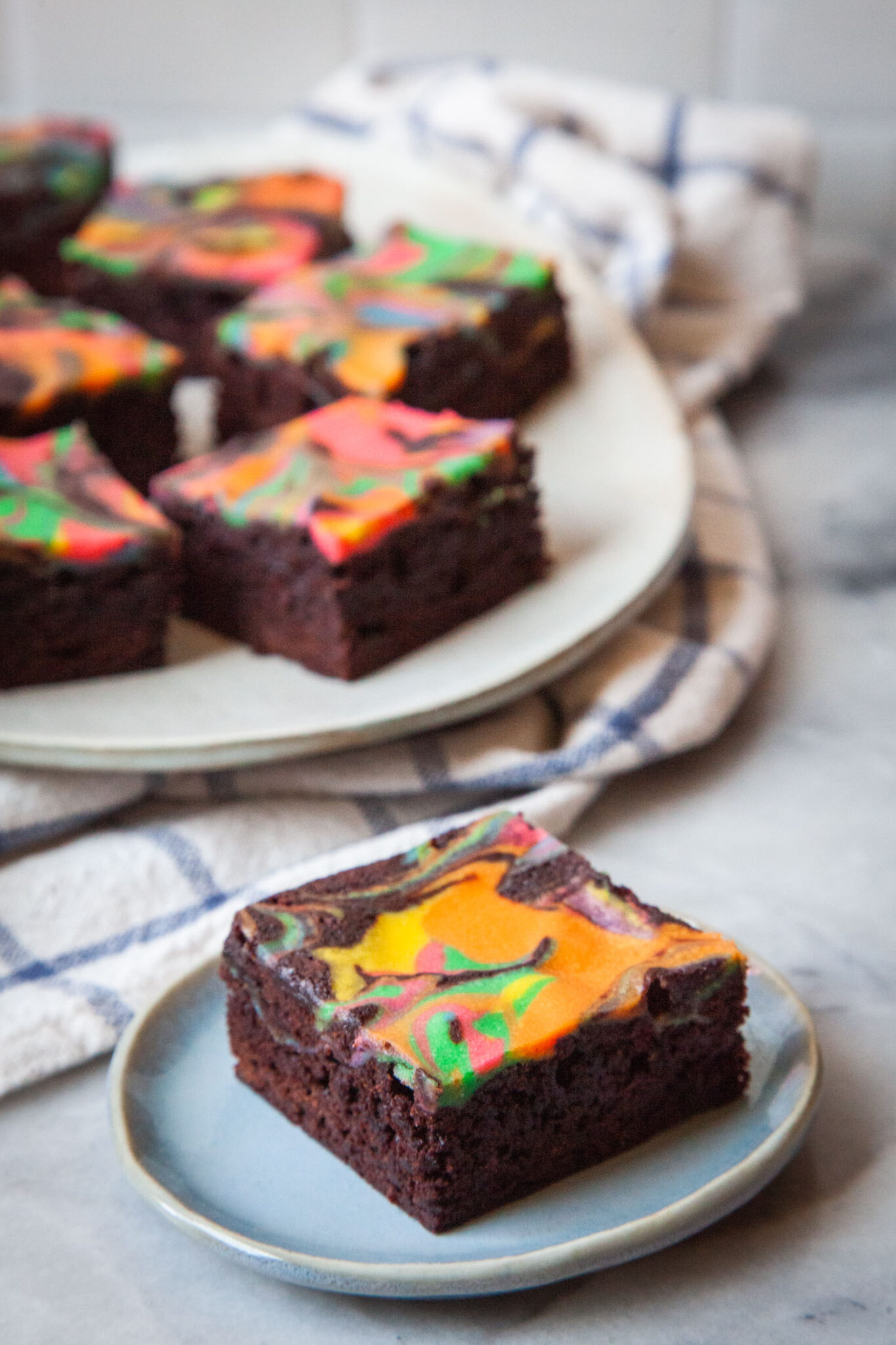 A single rainbow cheesecake brownie sitting on a small blue plate. There is an oval white plate sitting on a cloth napkin with more rainbow cheesecake brownies behind it.