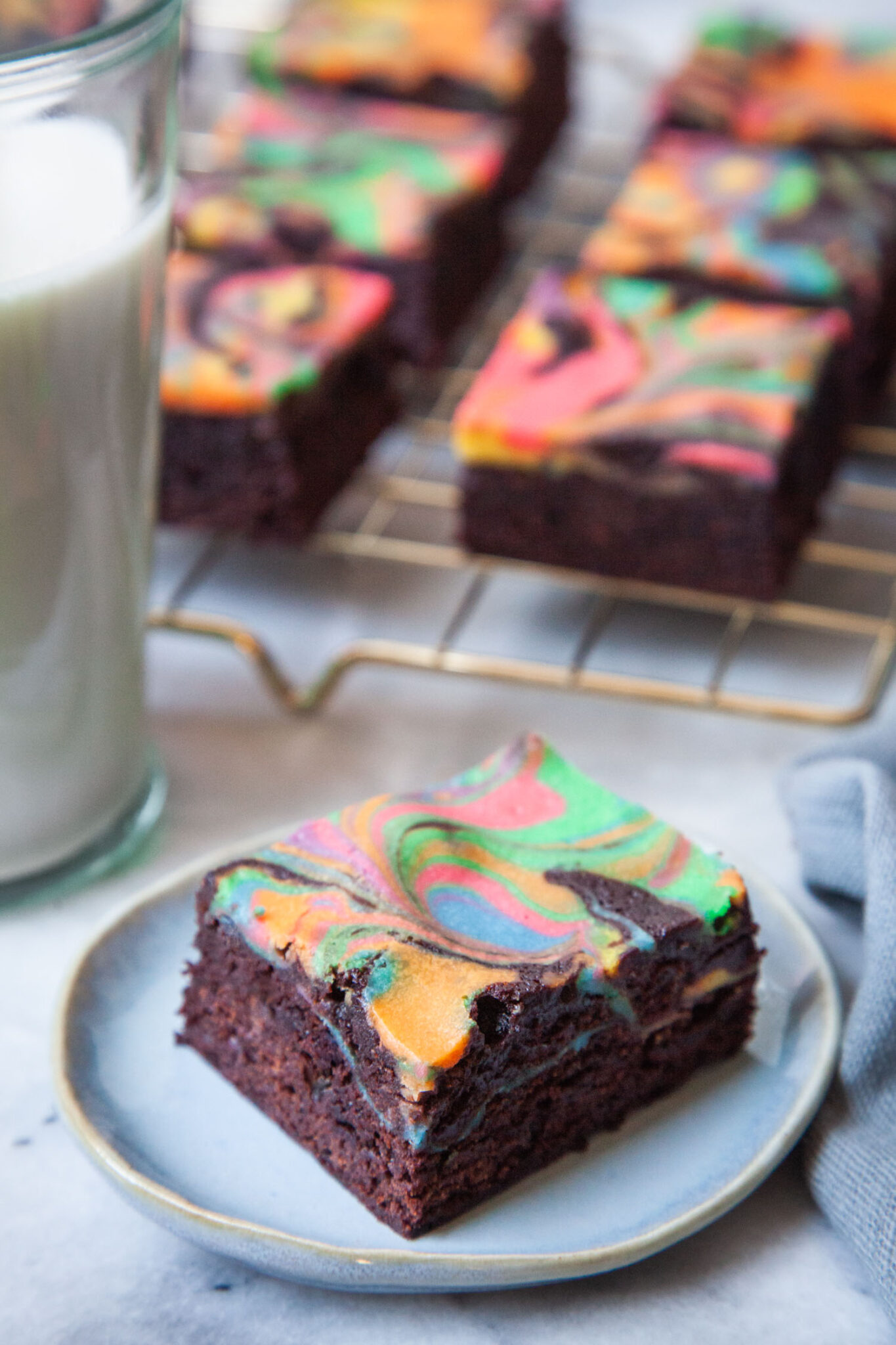 A single rainbow cheesecake brownie sitting on a small blue plate. There is a glass of milk next to it and a wire cooling rack of more rainbow cheesecake brownies behind it.