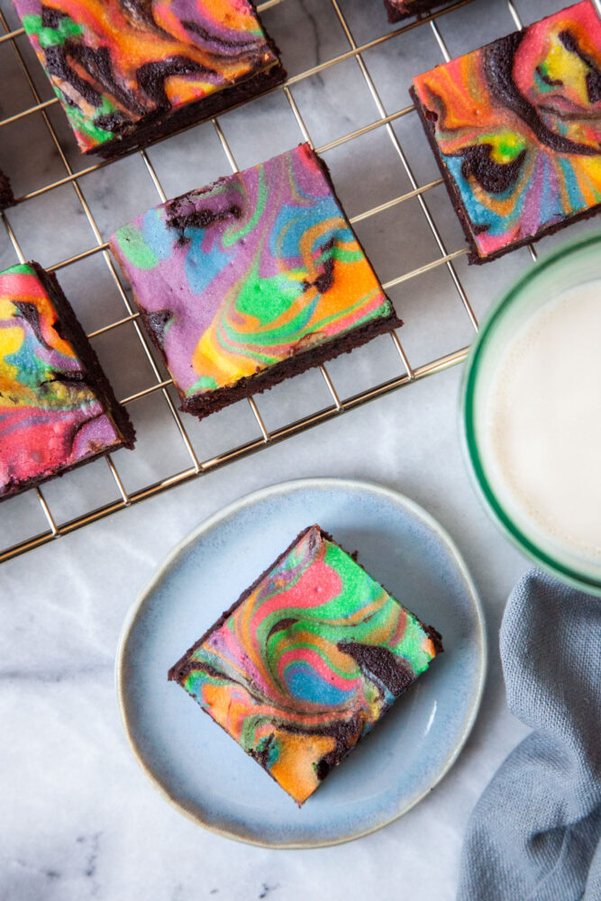 A single rainbow cheesecake brownie sitting on a small blue plate. There is a glass of milk and a wire cooling rack of more rainbow cheesecake brownies next to it.
