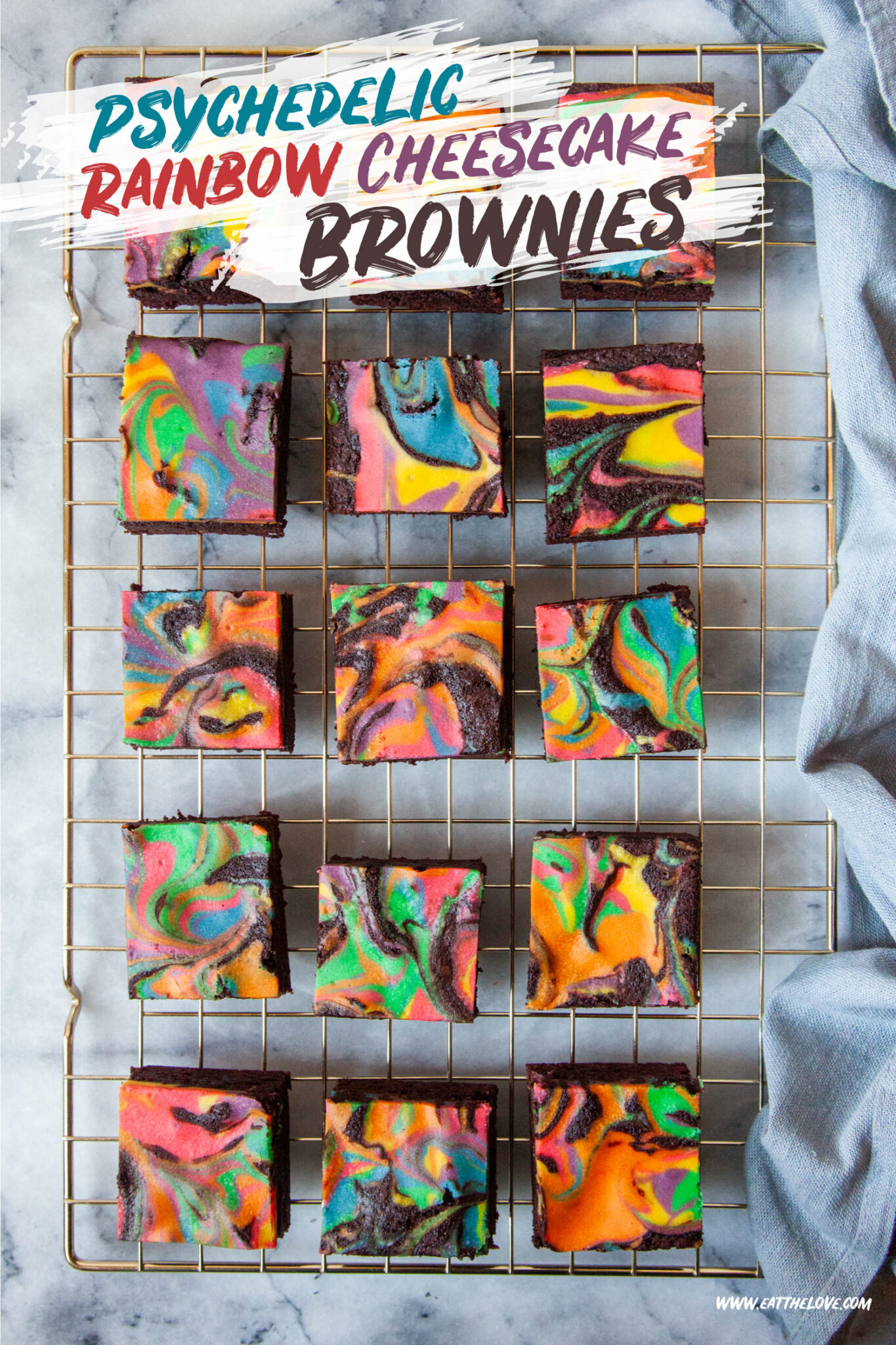 Rainbow cheesecake brownies sitting on a wire cooling rack. There is a blue gray cloth napkin next to the wire rack. 