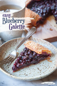 A slice of blueberry galette on a ceramic plate, with a fork next to it. There is a mug of tea behind it, along with the rest of the galette on a wooden cutting board.