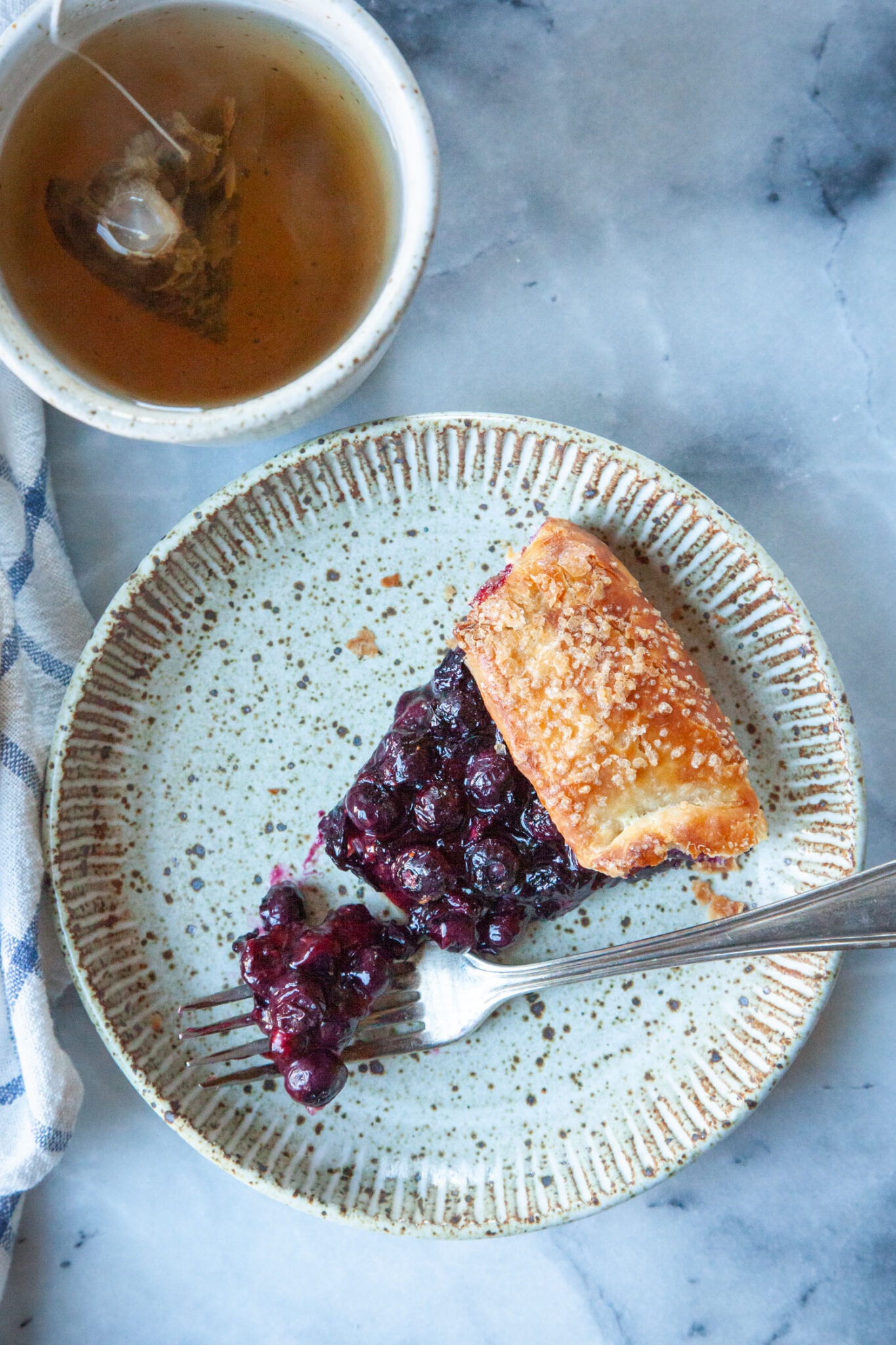 A slice of blueberry galette on a ceramic plate with a fork taking a piece out of the slice. There is a mug of tea and a cloth napkin next to the plate.