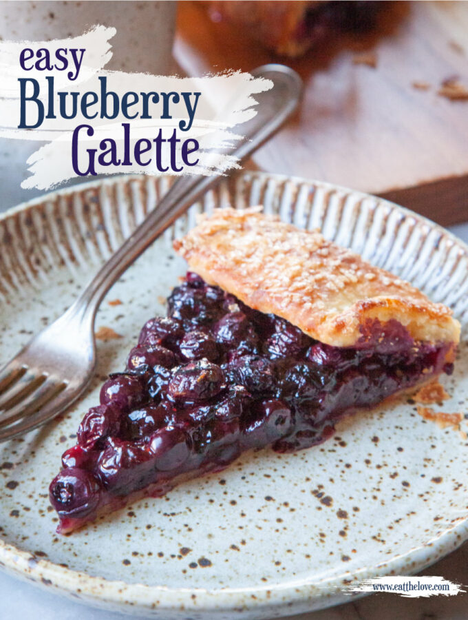 A slice of blueberry galette on a ceramic plate. There is a mug of tea behind it along with the remaining galette on a wooden cutting board. The title on the image says "Easy Blueberry Galette".