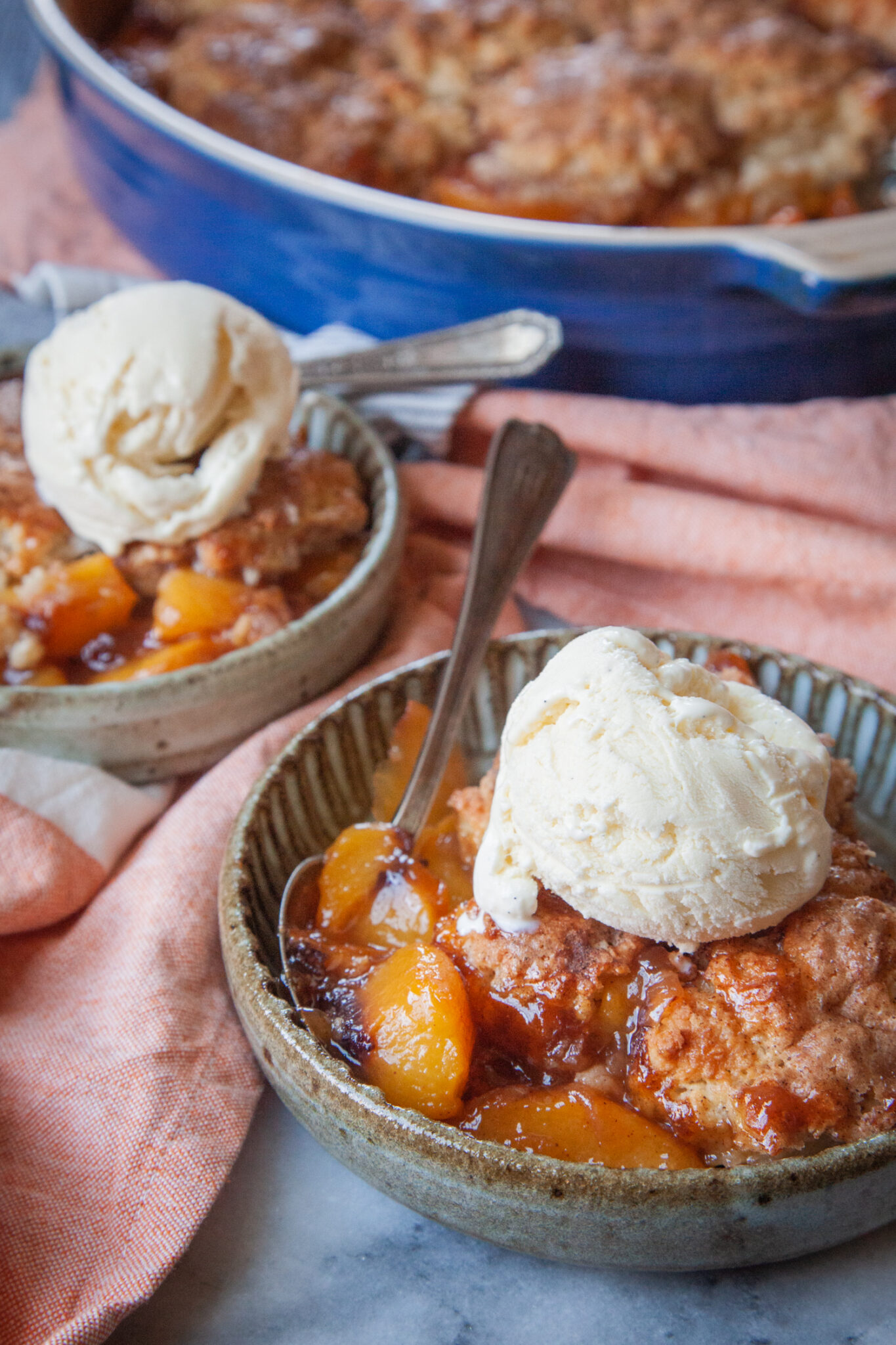 Two shallow bowls of peach cobbler with scoops of vanilla ice cream on top. The remaining cobbler is in a baking dish behind the bowls.