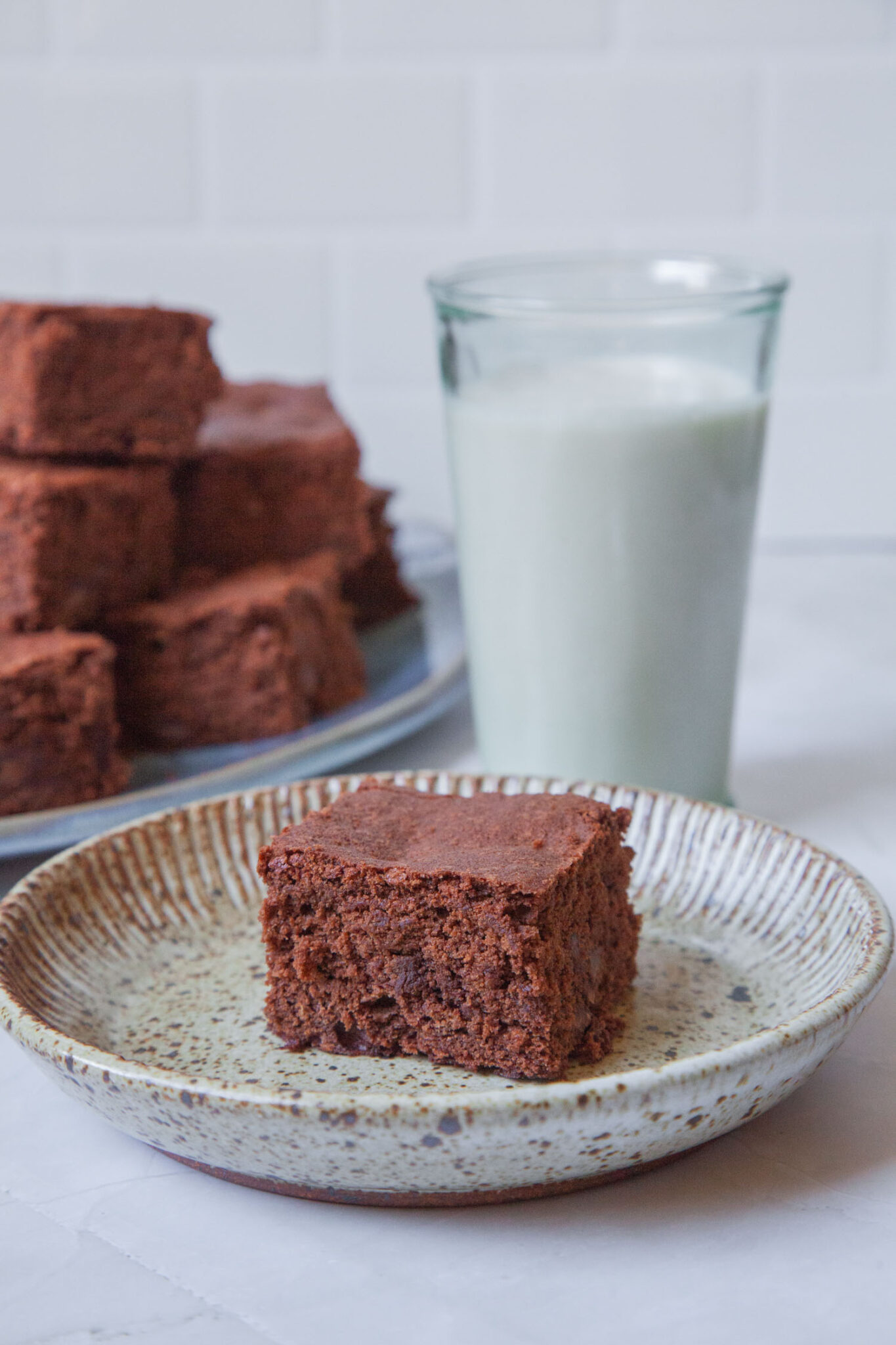 A cakey brownie sitting on a small plate, with a glass of milk and more brownies behind it.