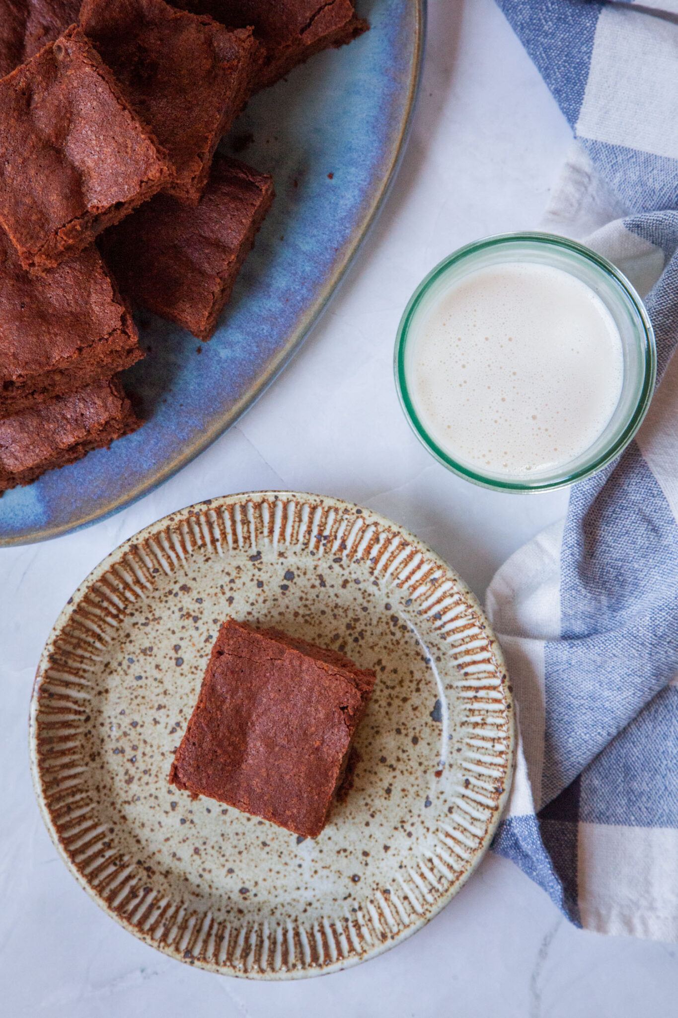 A cakey brownie on a small ceramic plate, with a glass of milk and more brownies next to it. There is a blue and white cloth napkin next to the plate.