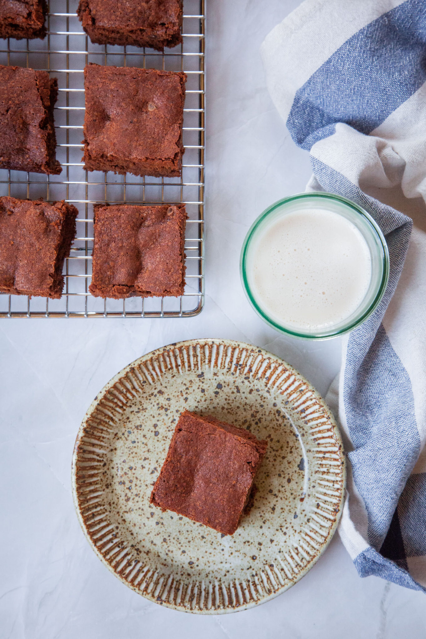 A cakey brownie on a small plate, with a glass of milk and more brownies on a wire cooling rack next to it. There is a blue and white cloth napkin next to the plate and glass of milk. 