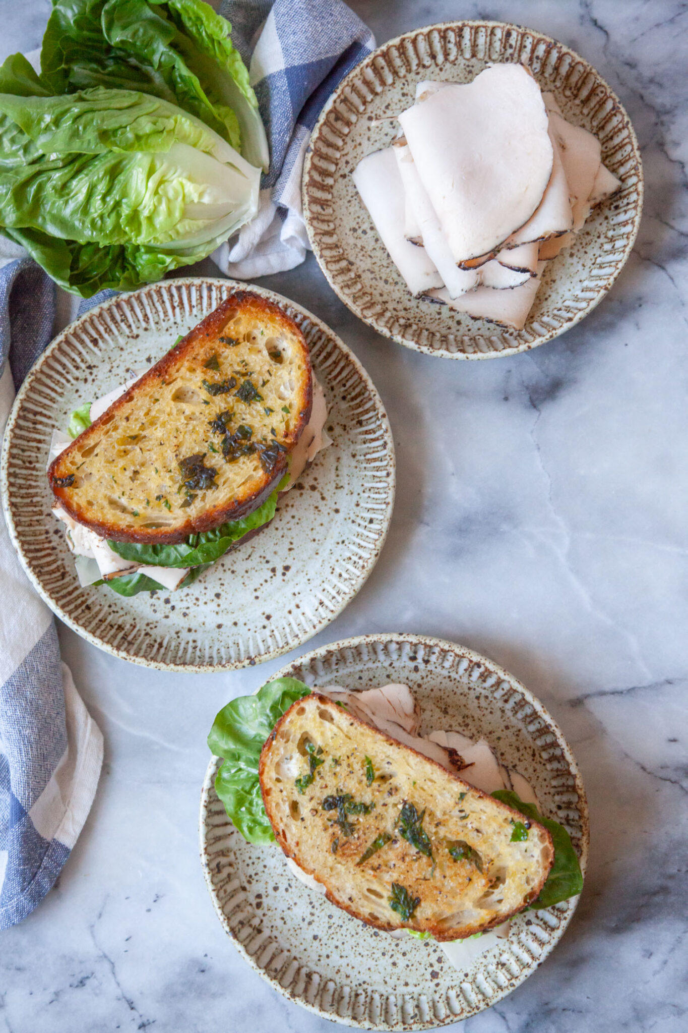 To chicken Caesar sandwiches on a ceramic plates with another plate of chicken deli meat next to them. There is a small head of Little Gem lettuce next to the plate with a cloth napkin.