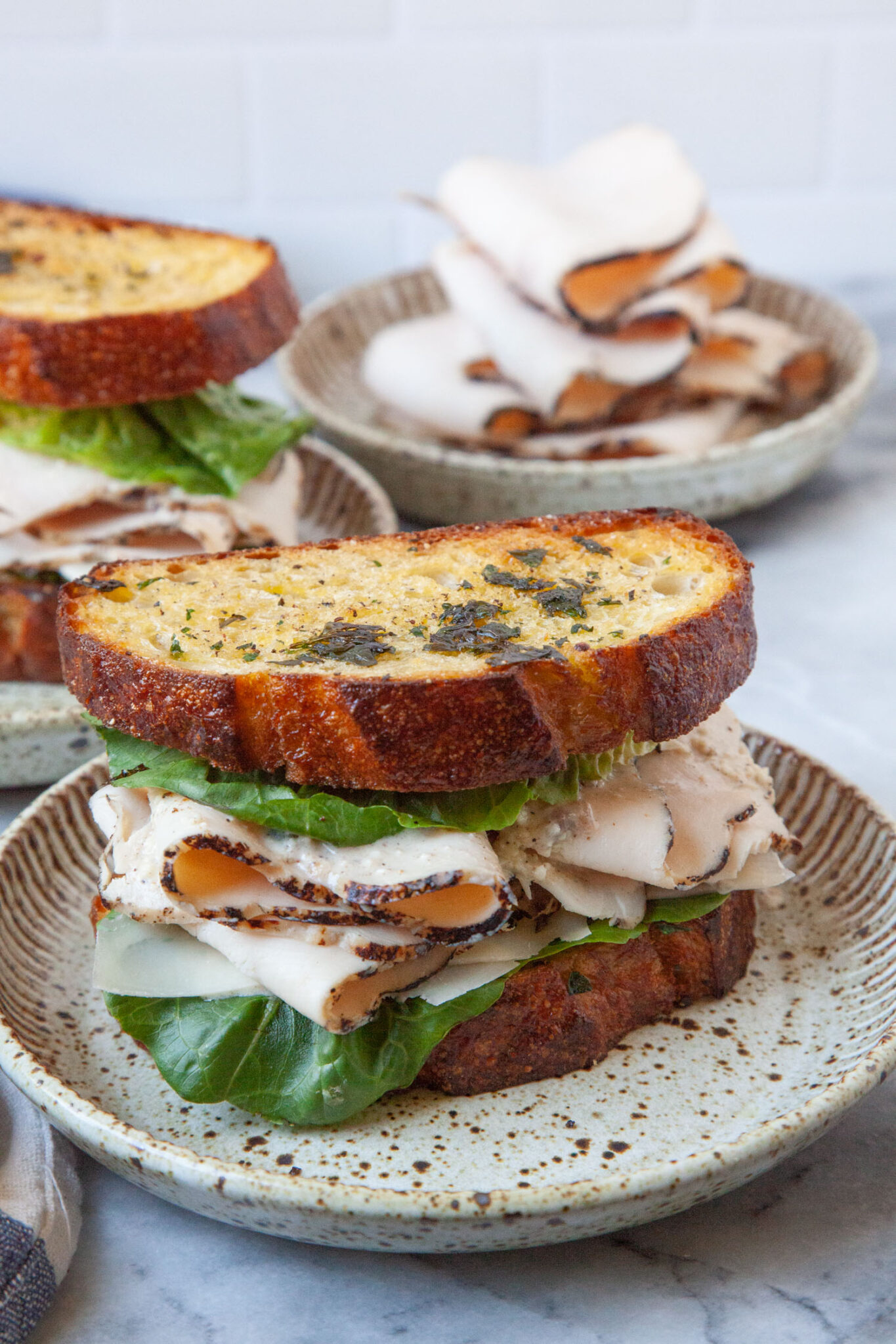 A chicken Caesar Sandwich on a plate, with another sandwich behind it. There is also a plate of sliced deli chicken behind both sandwiches.