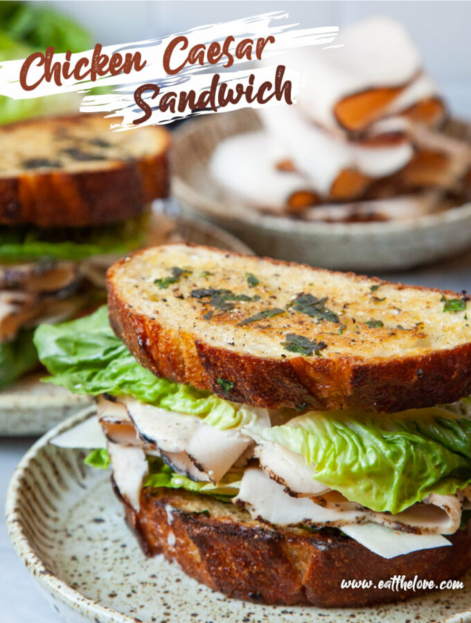 A chicken Caesar Sandwich on a plate, with another sandwich behind it. There is also a plate of sliced deli chicken behind both sandwiches. The text on the image says "Chicken Caesar Sandwich" as a headline, and in small print "www.eatthelove.com" in small print.