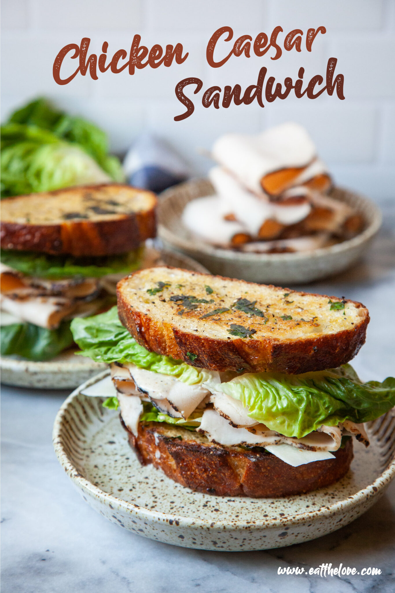 A chicken Caesar Sandwich on a plate, with another sandwich behind it. There is also a plate of sliced deli chicken behind both sandwiches. The text on the image says "Chicken Caesar Sandwich" as a headline, and in small print "www.eatthelove.com" in small print.