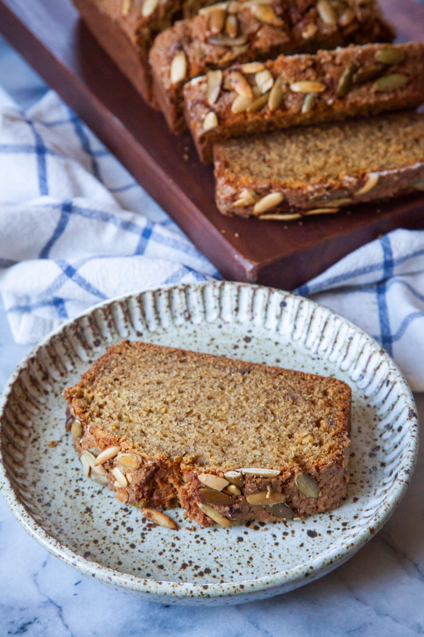 A slice of pumpkin banana bread on a ceramic plate, with the remaining pumpkin banana bread sliced on a cutting board behind the plate.