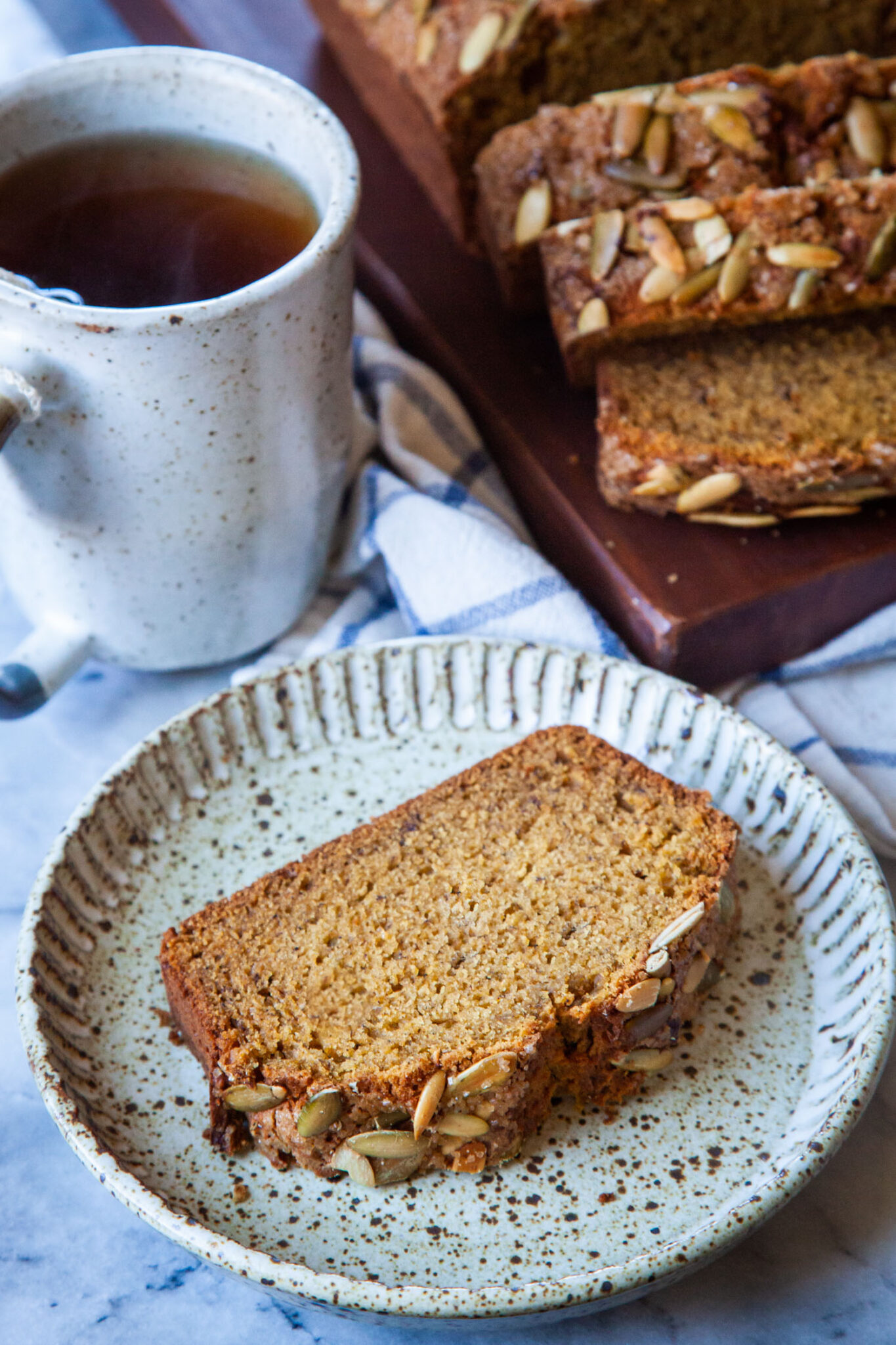 A slice of pumpkin banana bread on a ceramic plate, with a mug of tea and the remaining pumpkin banana bread sliced on a cutting board behind the plate.