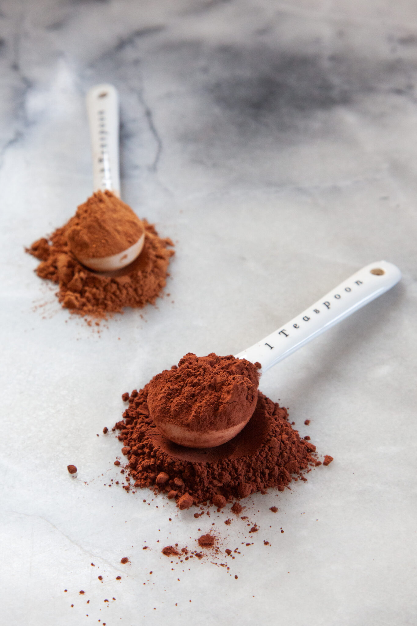 Two spoonfuls of different cocoa powder on a marble surface, with darker Dutch-processed cocoa in front, and natural cocoa powder in back.