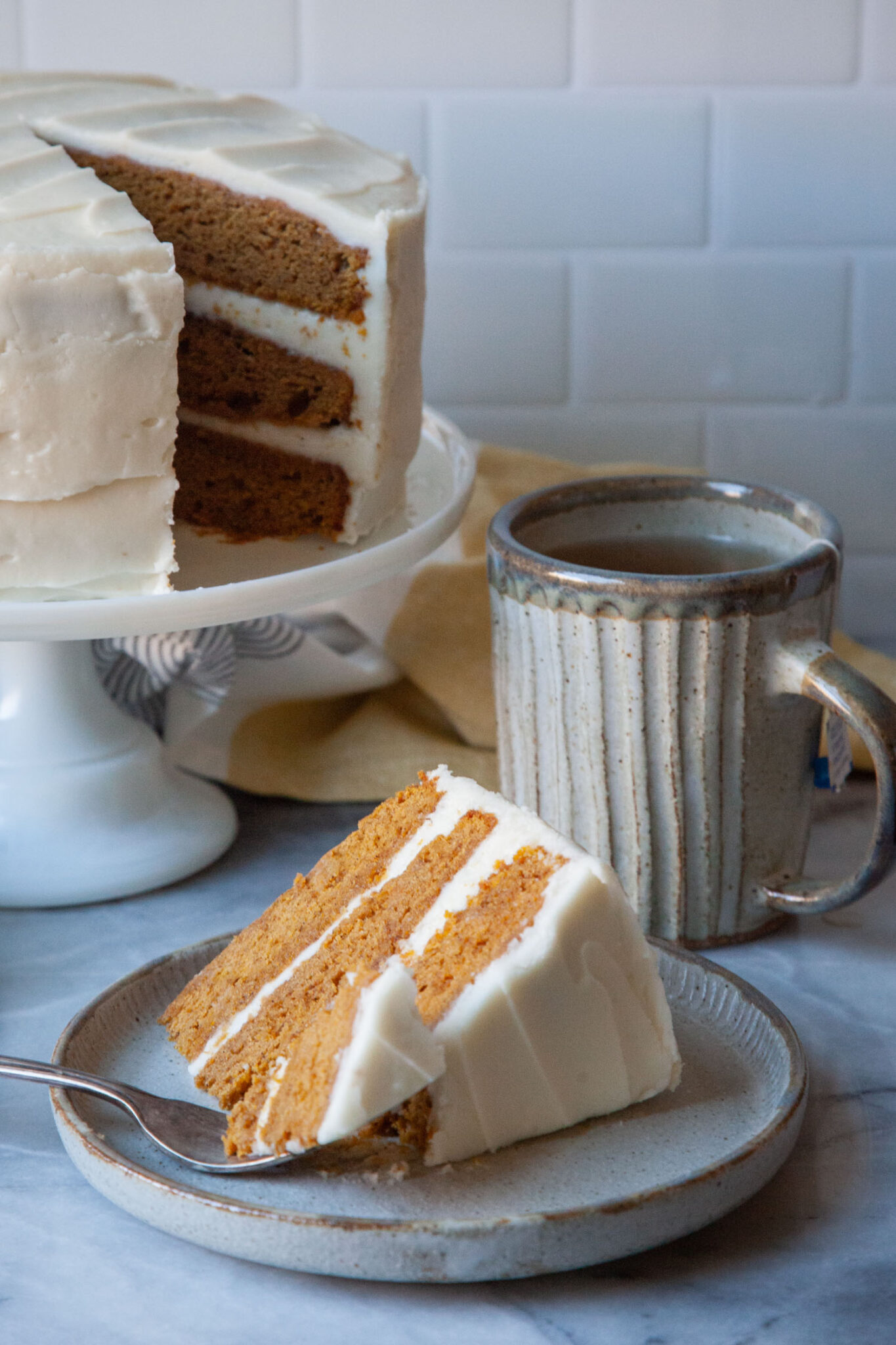 A slice of pumpkin layer cake on a plate, with a fork that has taken a bite size piece out of the slice. Behind the plate is the remaining cake on a cake stand and a mug of tea.