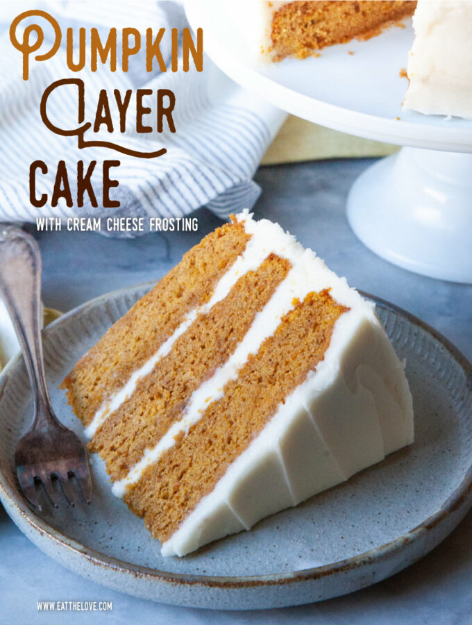 A slice of pumpkin layer cake with cream cheese frosting on a plate, with the remaining cake on a cake stand behind it.