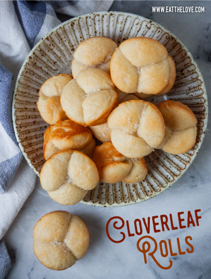 A bowl of cloverleaf shaped bread rolls, with one roll on the counter. A blue and white cloth napkin is next to the bowl. The type on the image says Cloverleaf Rolls.